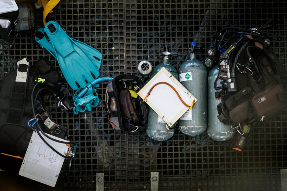 A collection of scuba gear, air tanks, fins and a dive slate on the deck of a pier