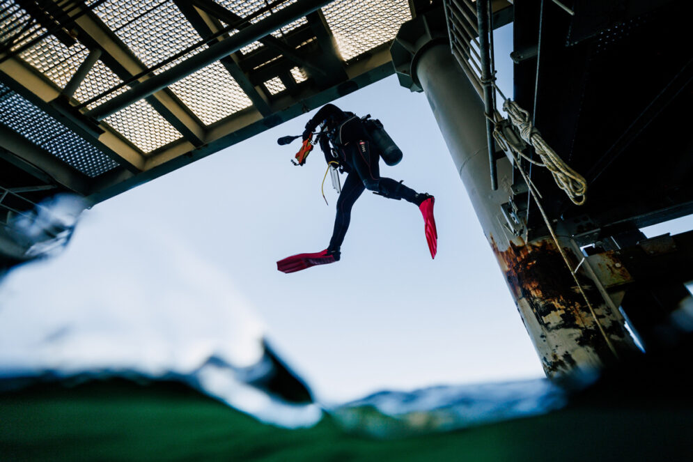 A scuba diver wearing a black wetsuit and red fins jumps into the water