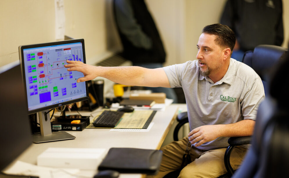 A man in a polo shirt points to a monitor showing a colorful data display.