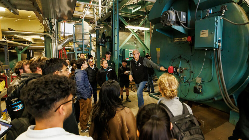 In a large warehouse-like room, a professor surrounded by students gestures to several large green tanks