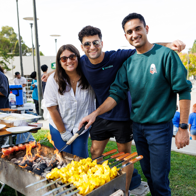 Three people smile near a grill with food on kebabs