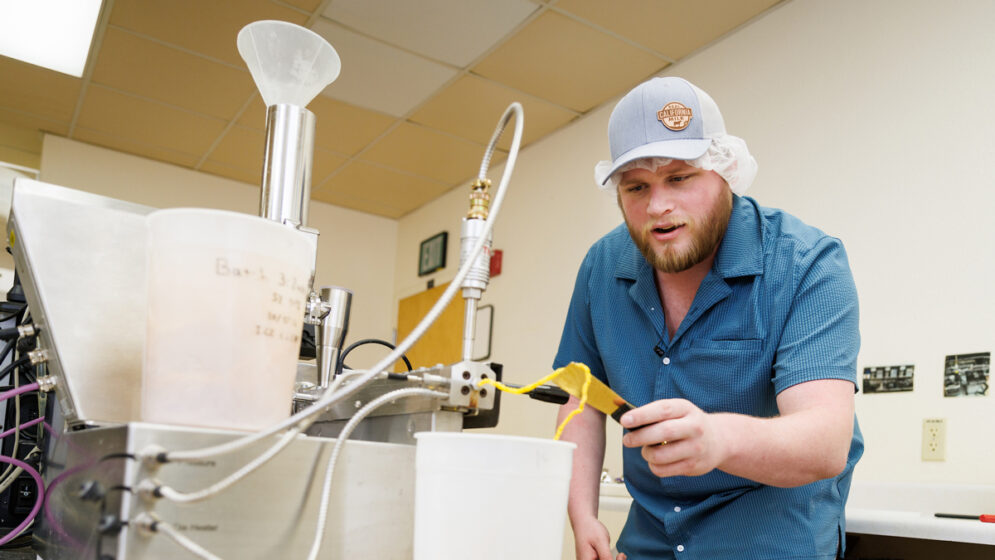 A student wearing a hat and a hair net works with an extruder in a culinary lab