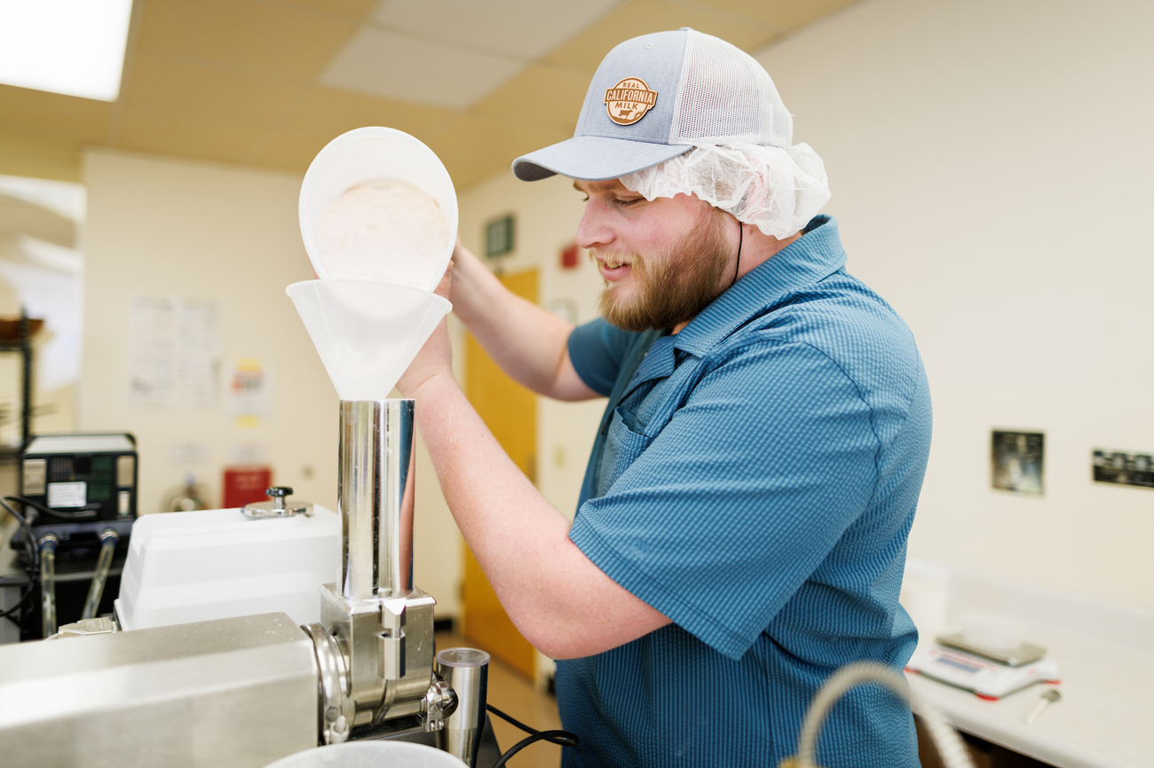 A food science student wearing a hairnet and a hat pours dry ingredients into an extruder machine in a culinary lab