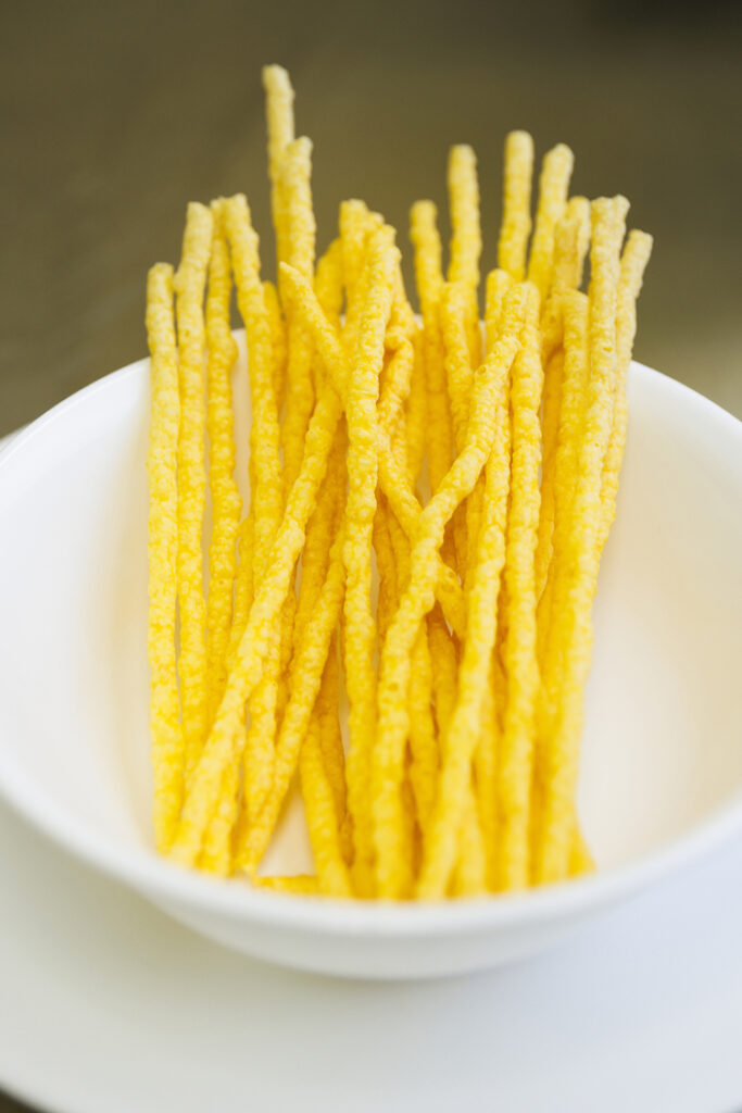 A white bowl filled with yellow rods of carrot-based crunchy snacks