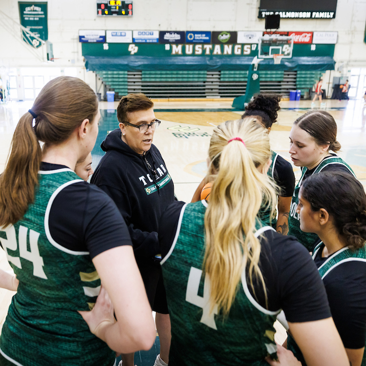 A coach speaks to women's basketball players in a huddle on the court