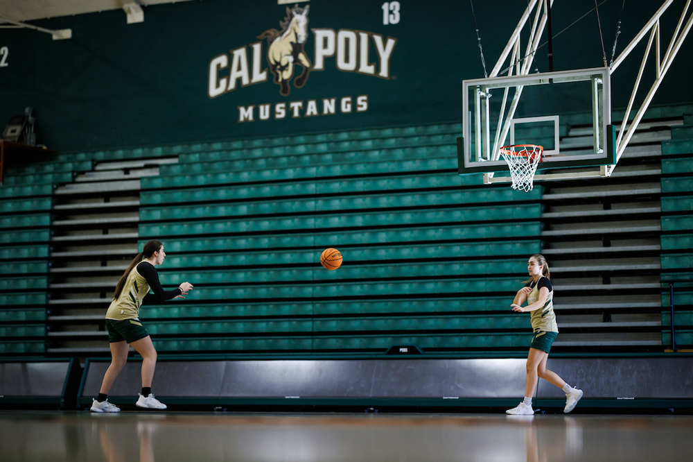 Two members of the women's basketball team pass a basketball near a Cal Poly Athletics logo