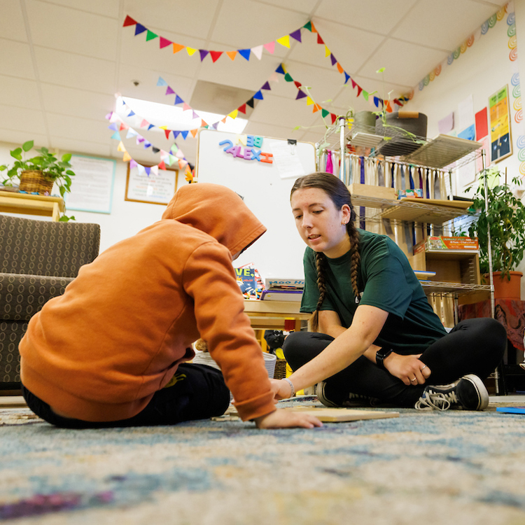 A student teacher helps a young child read a book on the floor of a daycare center