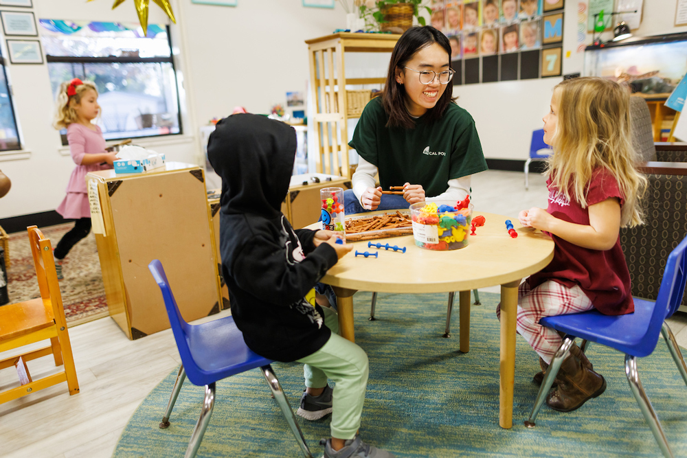 A student teacher sits at a small table with two young children to play with toys at a childcare center