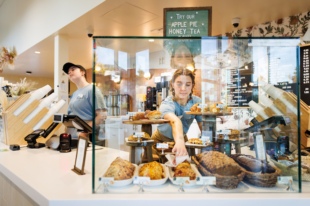 Two student employees work at the front counter of a coffee shop near a display of pastries