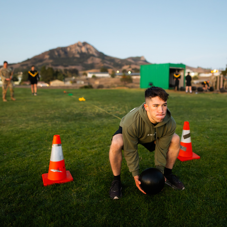 A person squats to lift a black medicine ball between two orange cones on grass in front of Bishop Peak