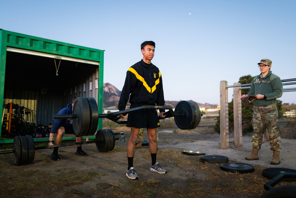 Two people lift barbells while a leader in fatigues holds a clipboard