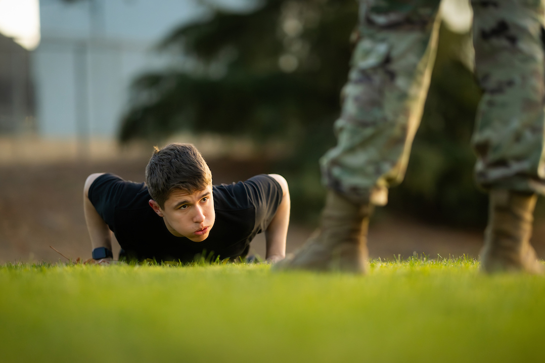 An ROTC cadet performs an exercise on their stomach while looking toward a standing military officer in fatigues and boots