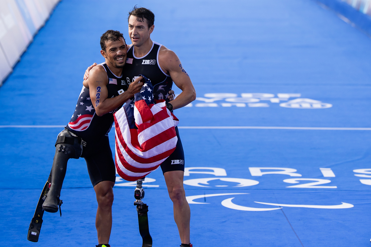 Paralympic triathletes Mark Barr and Mohamed Lahna cross the finish line holding an American flag