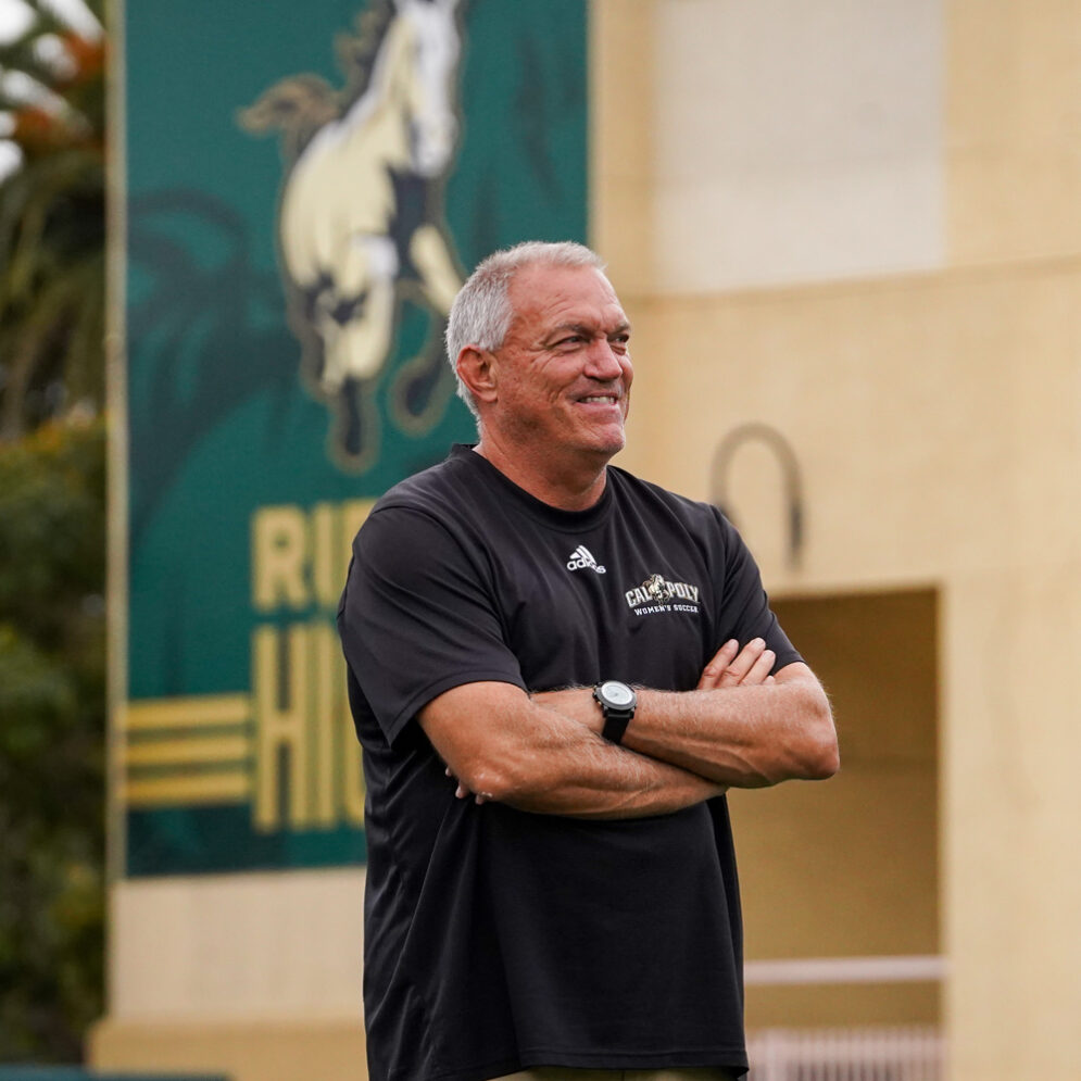 Coach Alex Crozier wears a black Cal Poly shirt while smiling in front of a Cal Poly Athletics banner in Spanos Stadium