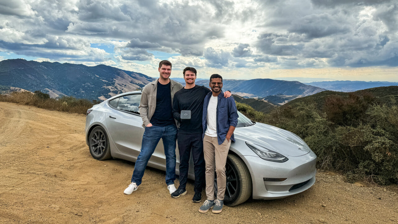 Three members of the NeoCharge team smile standing next to a silver Tesla overlooking mountains