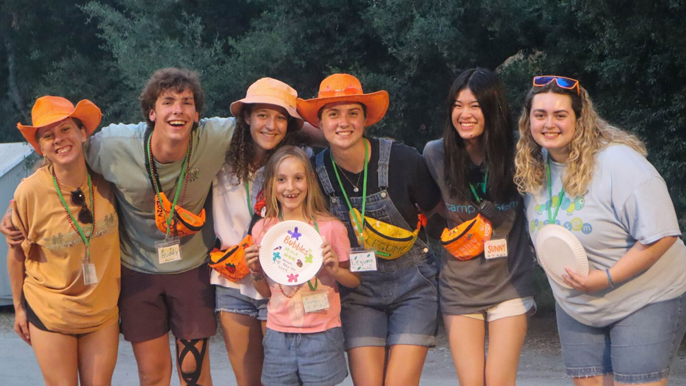 A group of young people stands with children at a summer camp