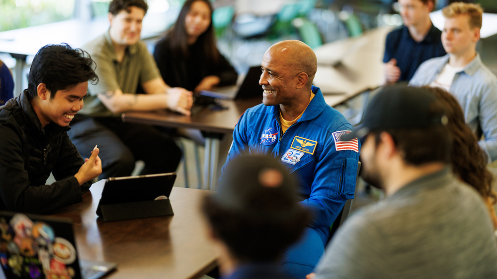 Astronaut Victor Glover wears a blue flight suit while sitting in a classroom talking to college students