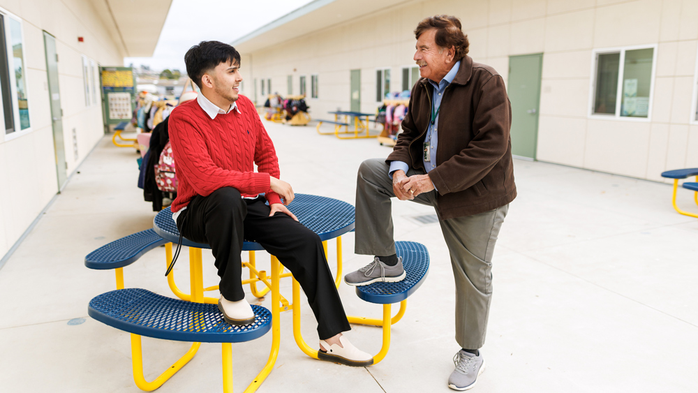 A student and a school principal sit talking at a school lunch table