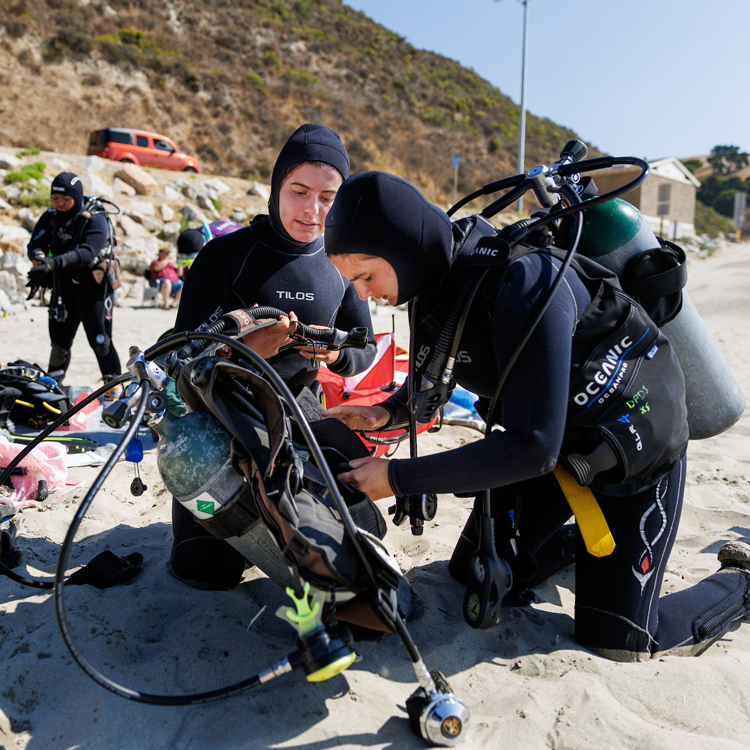 Three divers in black wet suits check their scuba gear and air tanks on a sandy beach