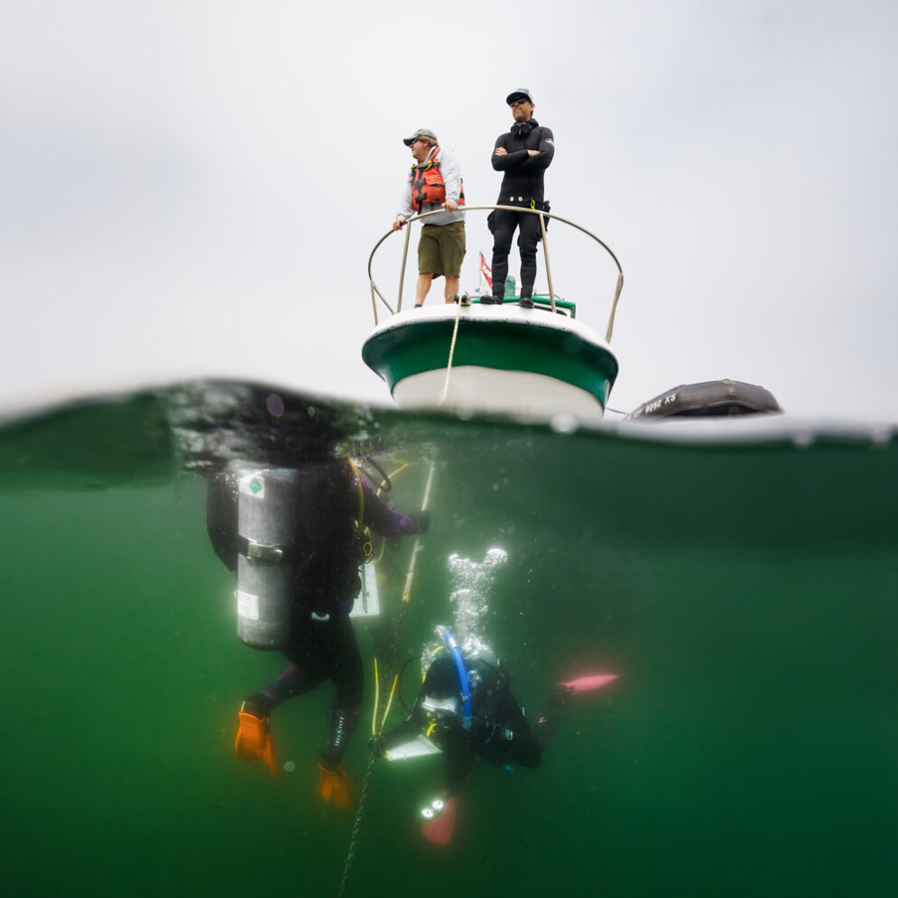 Two people stand on the bow of a boat while two scuba divers hold an anchor line below the surface of the water