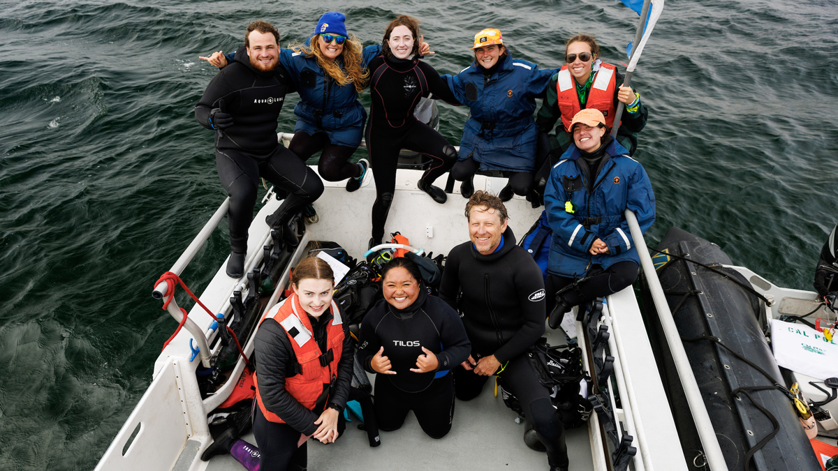 Nine scuba divers in wetsuits and jackets smile while sitting on the back of a boat in the ocean