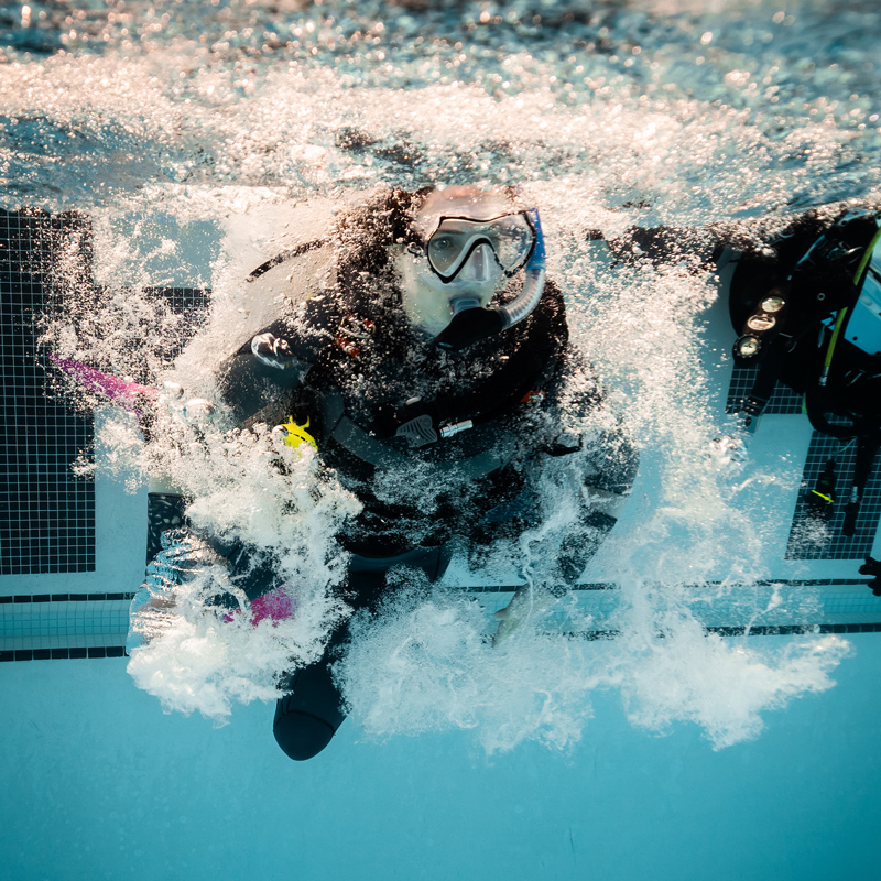 A student scuba diver jumps into the a swimming pool wearing scuba gear, creating a cloud of bubbles