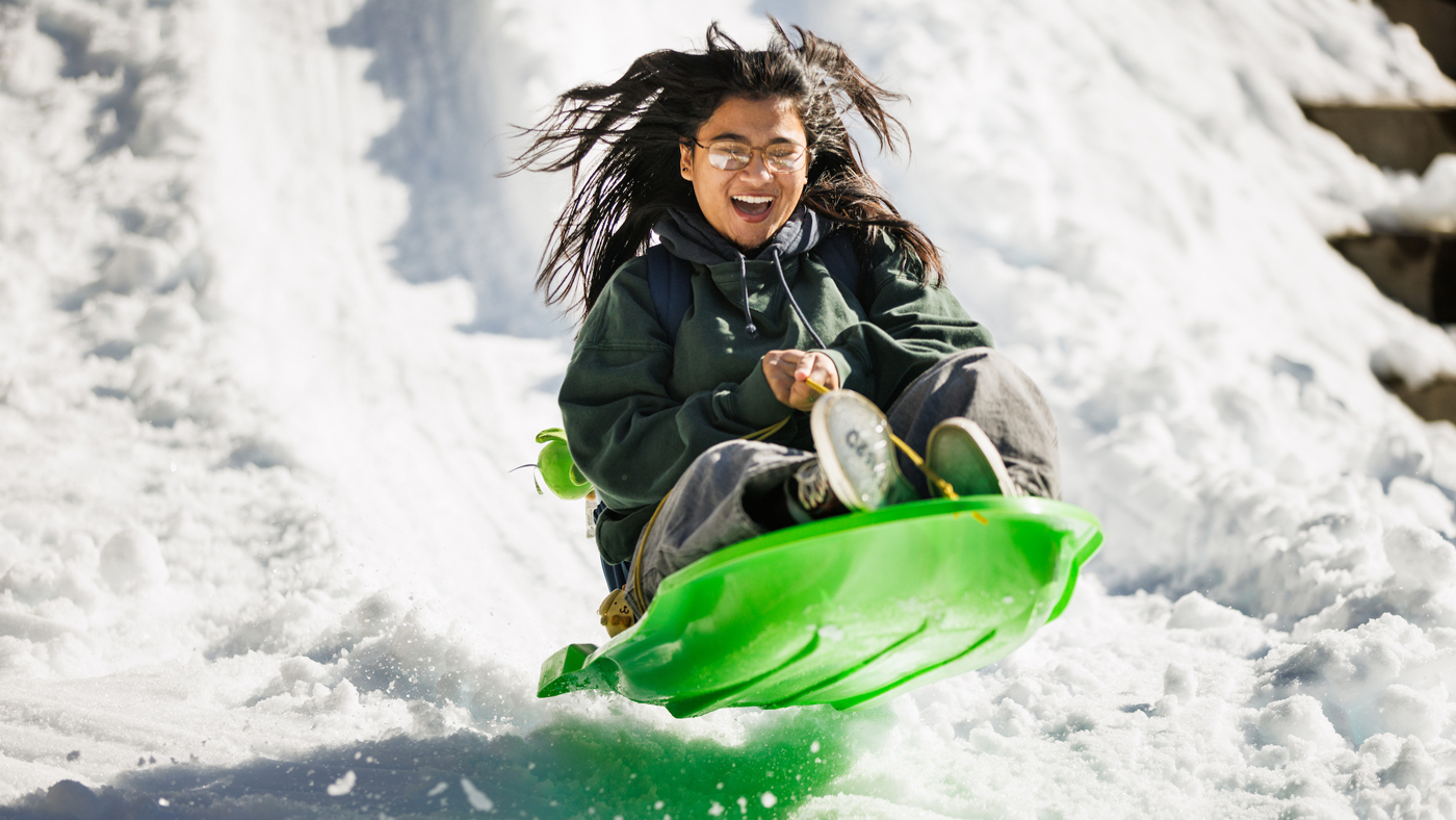 A smiling student riding a green, plastic sled, slides down a hill of snow