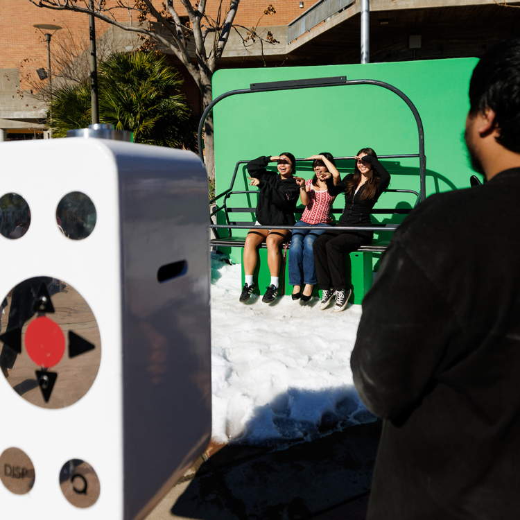 Three students sit on a ski lift in front of a green screen with snow at their feet.