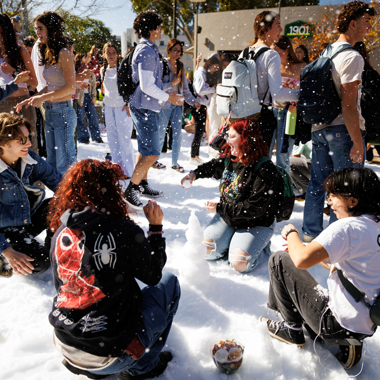 Student kneel around a small snowman in snow as falls in Cal Poly's University Union Plaza