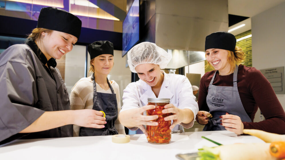 Four students in aprons and hair coverings surround a jar of kimchi in a culinary lab