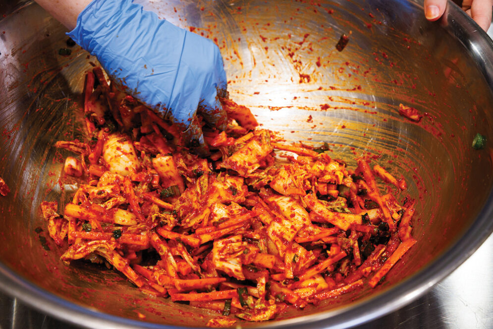 A student wearing a blue glove mixes kimchi ingredients in a metal bowl