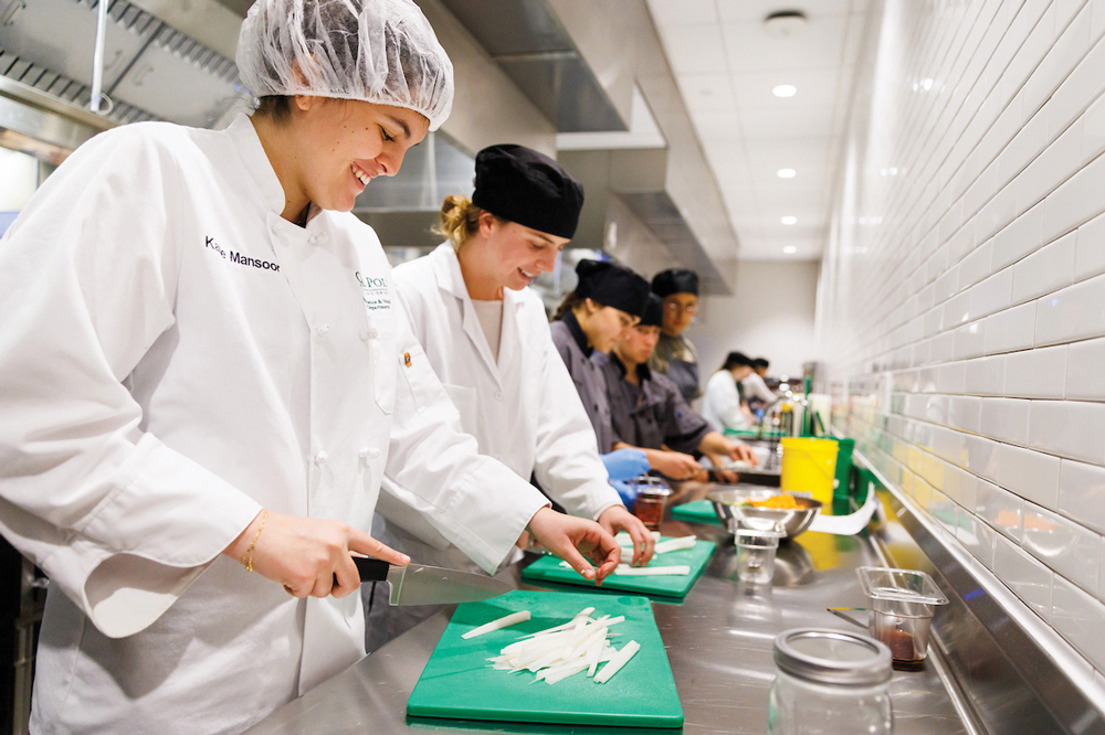 Students in hair nets and chef's coats chop vegetables on a steel countertop