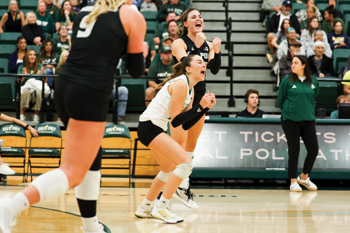 Cal Poly women's volleyball players celebrate on the court as a coach and sideline commentator look on