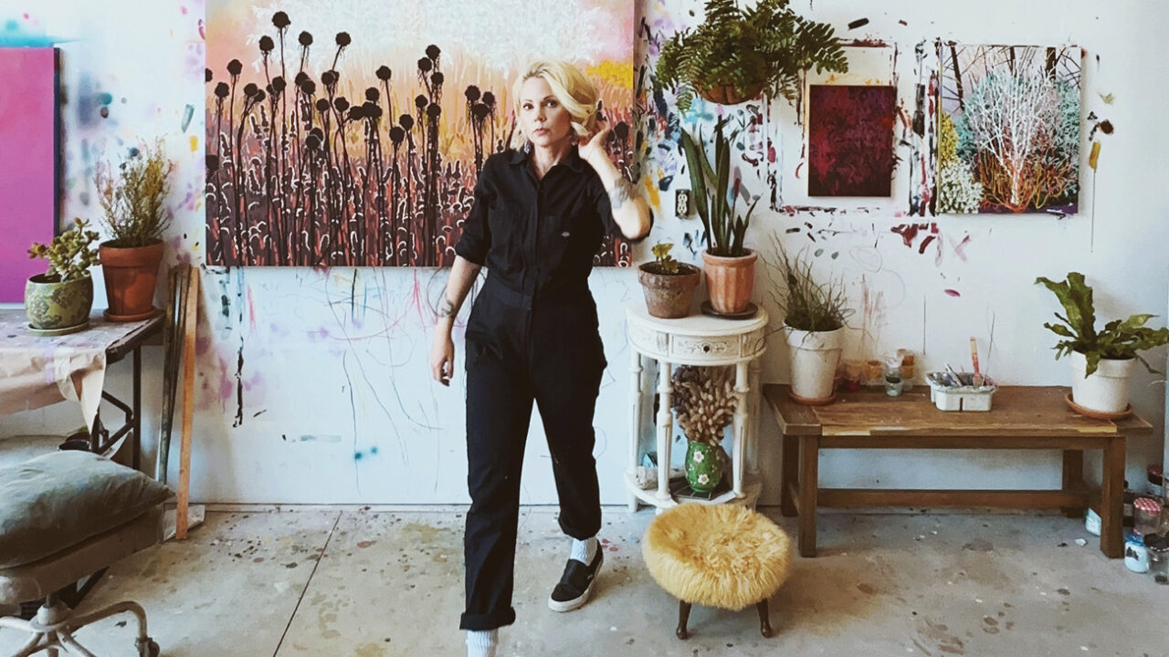 Professor Sarah Frantz in a black jumpsuit in her art studio, featuring paintings on the walls and eclectic furniture