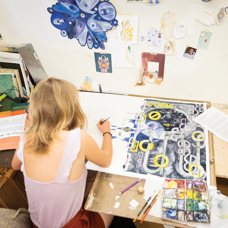 A student leans over a desk to paint a piece of art