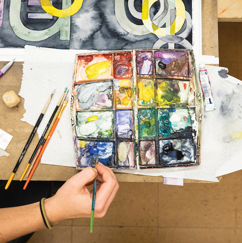 An artist dips a paint brush into a well-used palette with rainbow colors