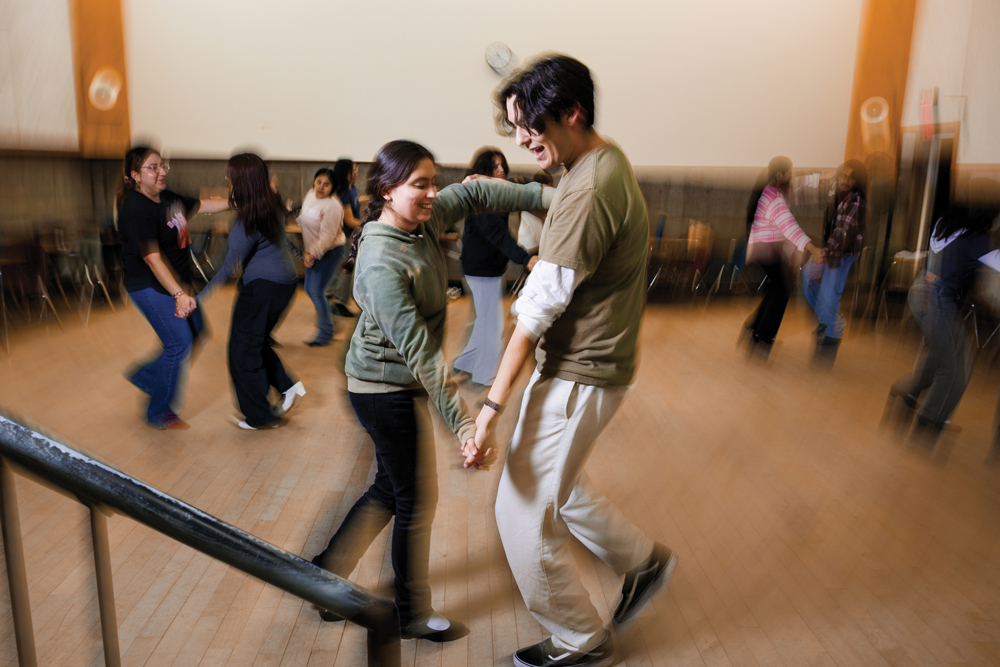 Two students perform a traditional Mexican folk dance among pairs of other dancers on a hardwood floor
