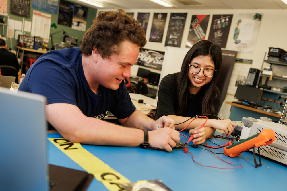 Two engineering students test a circuit board with tools in a lab