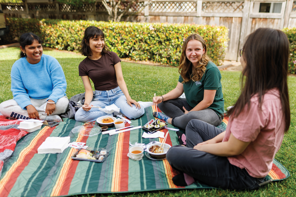 Four students sit on a colorful blanket eating food with chopsticks