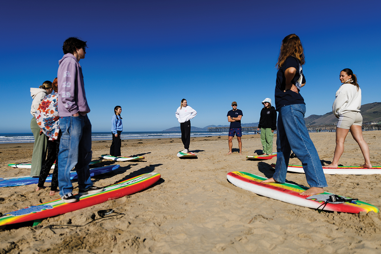 On the sand of a sunny beach, a group of young people stand in a circle on foam surfboards.