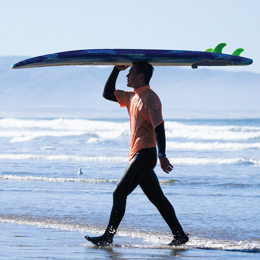A young man in a wetsuit and orange jersey carries a surfboard on his head as he walks out of the ocean.