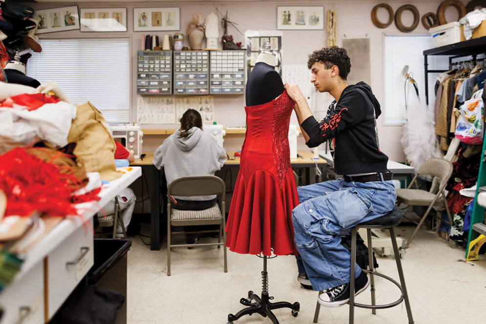 A young man in a black hoodie sits in front of a tailor's mannequin, hand stitching a red dress.