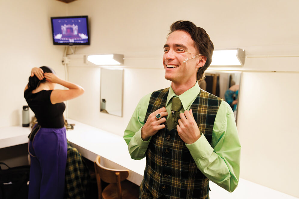 A young man in a green shirt and tie and plaid vest adjusts his collar in a dressing room.