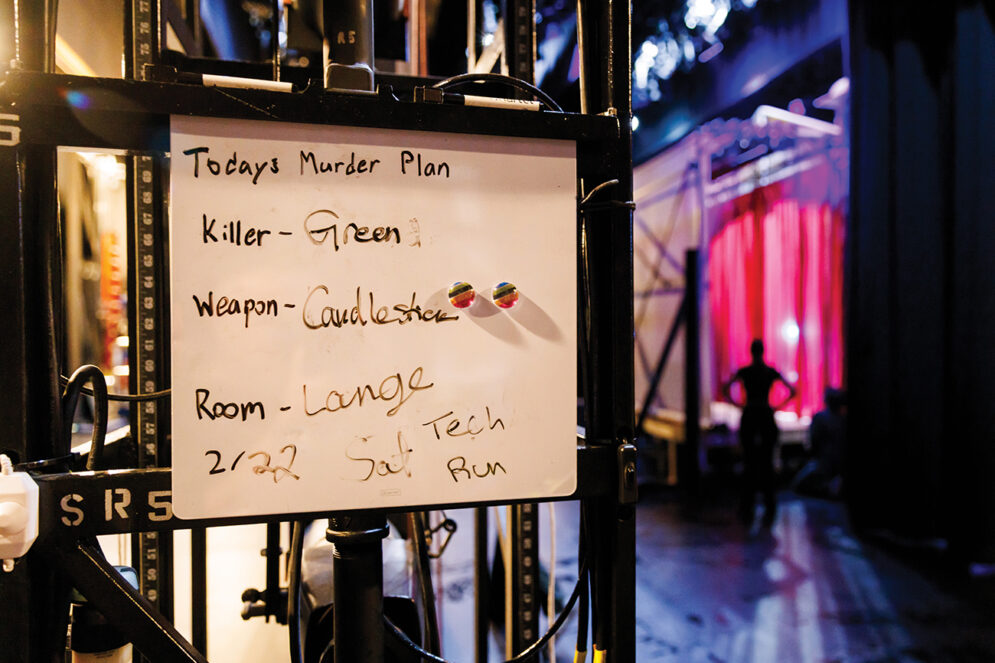 A dry erase whiteboard reading "Today's Murder Plan: Killer - Mr. Green, Weapon: Candlestick, Room: Lounge" hangs on a rack, with a backlit red curtain in the background.