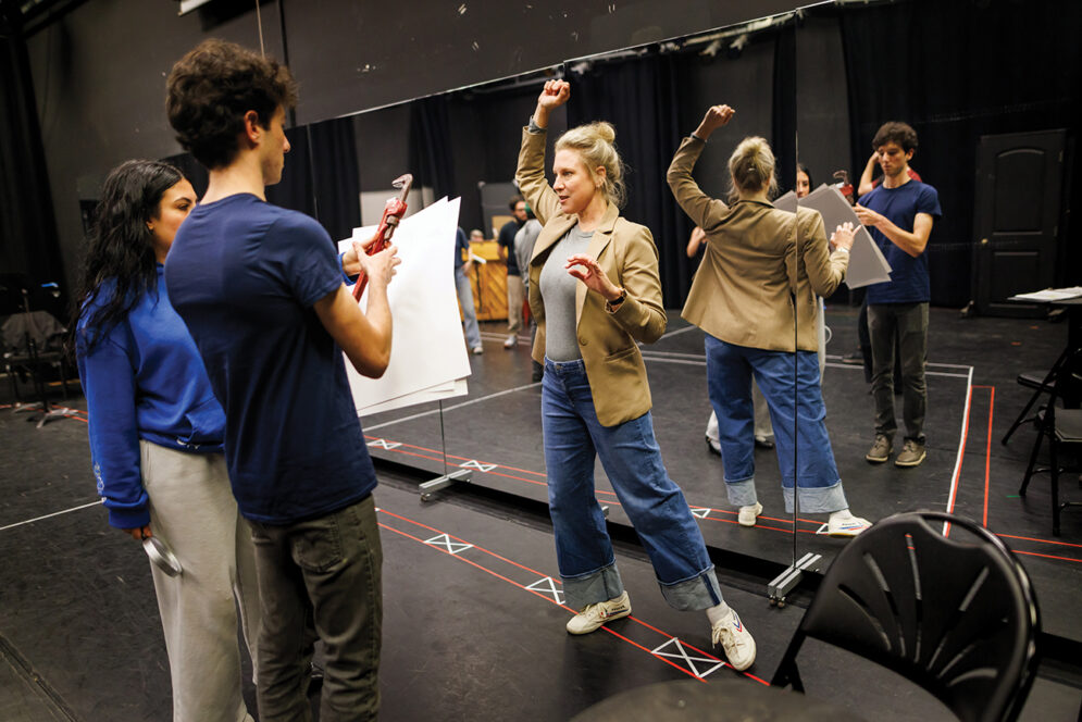 In front of a panel of large mirrors, a woman in a blazer demonstrates a dramatic gesture to a young man and young woman.