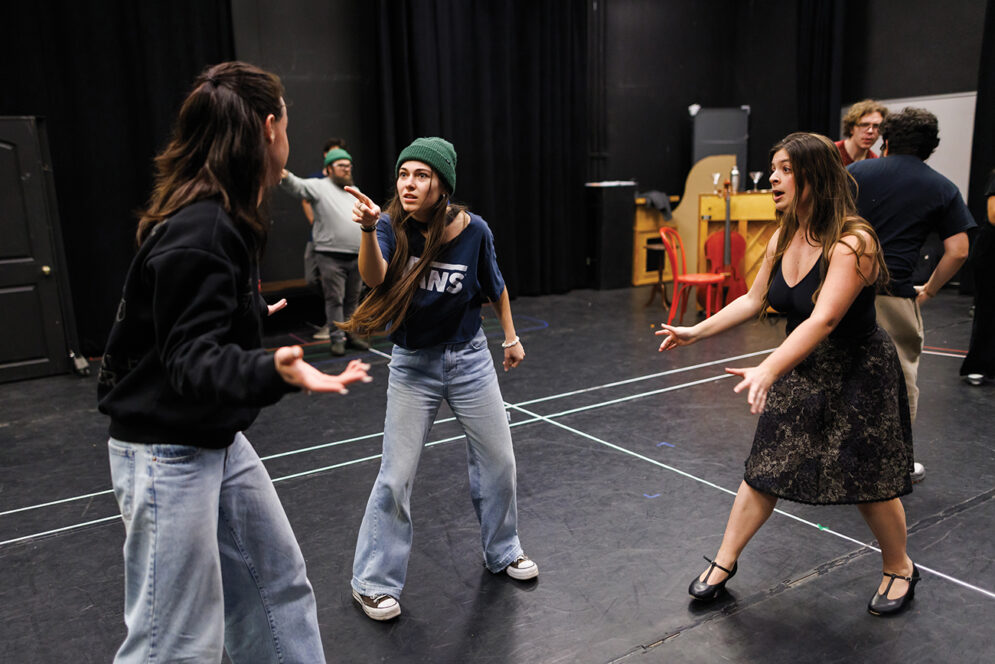 Two young women gesture dramatically at a young man in a room painted all black.