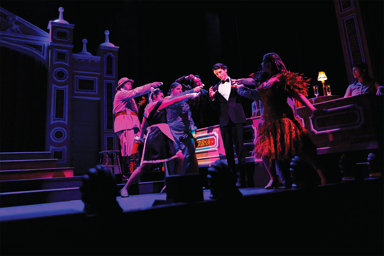 On a pink-lit stage, a group of flashily-dressed characters point menacingly at a young man in a tuxedo.