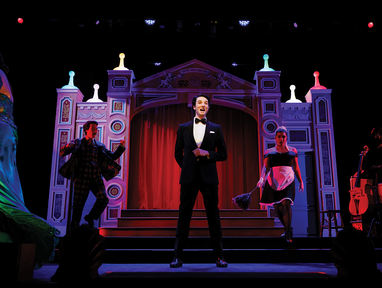 A young man in a tuxedo sings on stage in front of an elaborate backdrop, while a man in a suit an a suit and a woman in a maid's uniform dance nearby.