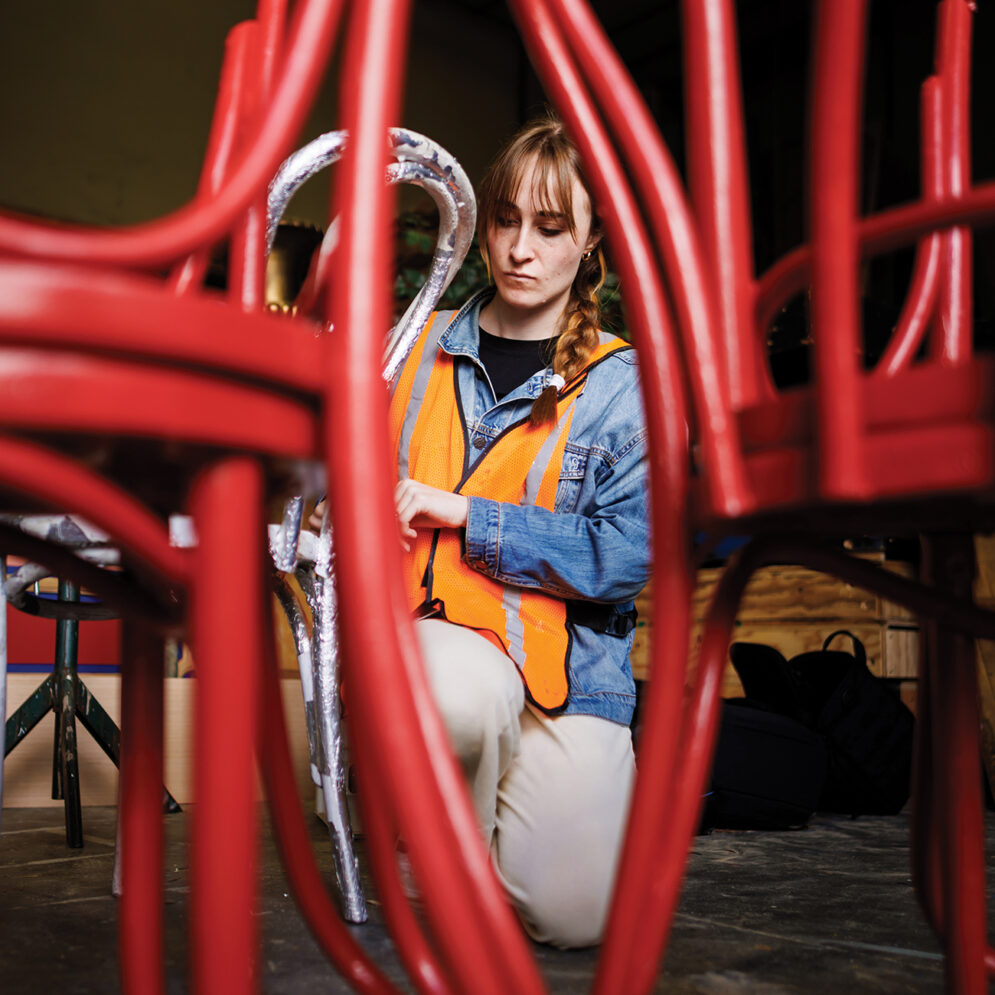 A young woman in a bright orange vest kneels while painting one of several stacked chairs.