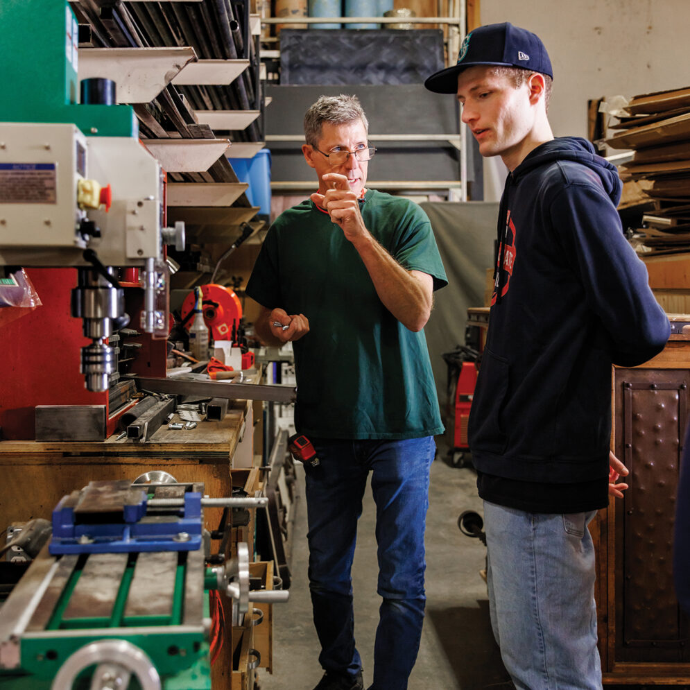 An older man gestures while talking to a young man in a cap in front of a workbench covered with a variety of power tools.